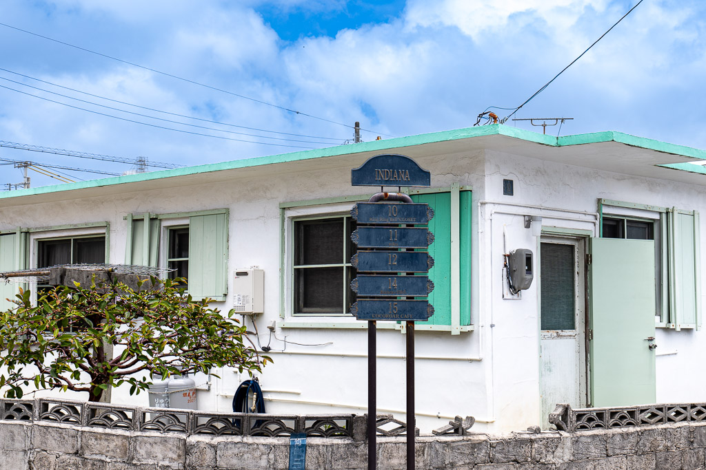 港川 INDIANA block — white walls with mint shutters and shop signboard, Okinawa Urasoe American Stateside housing signature shot