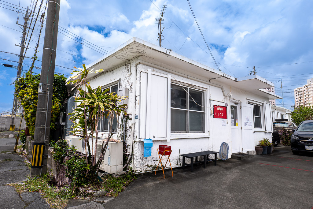 港川 (Minatogawa) white exterior with red box and chair, alley mood — Okinawa Urasoe street