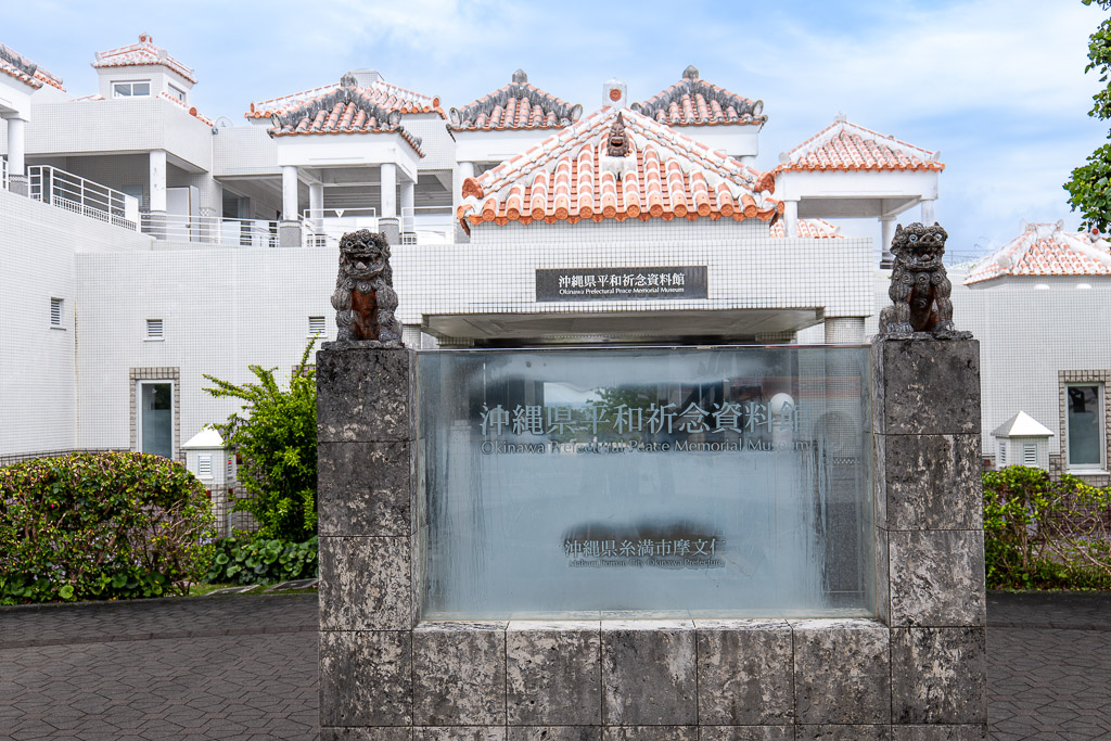 Glass nameplate and shisa lions at Okinawa Prefectural Peace Memorial Museum — white walls and red Ryukyu tiles