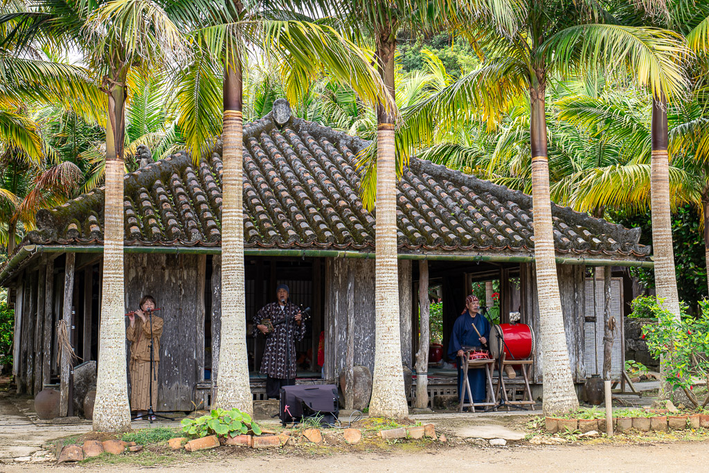 Traditional kominka exterior with palm trees, red-tile roof, and staff in traditional Ryukyuan dress