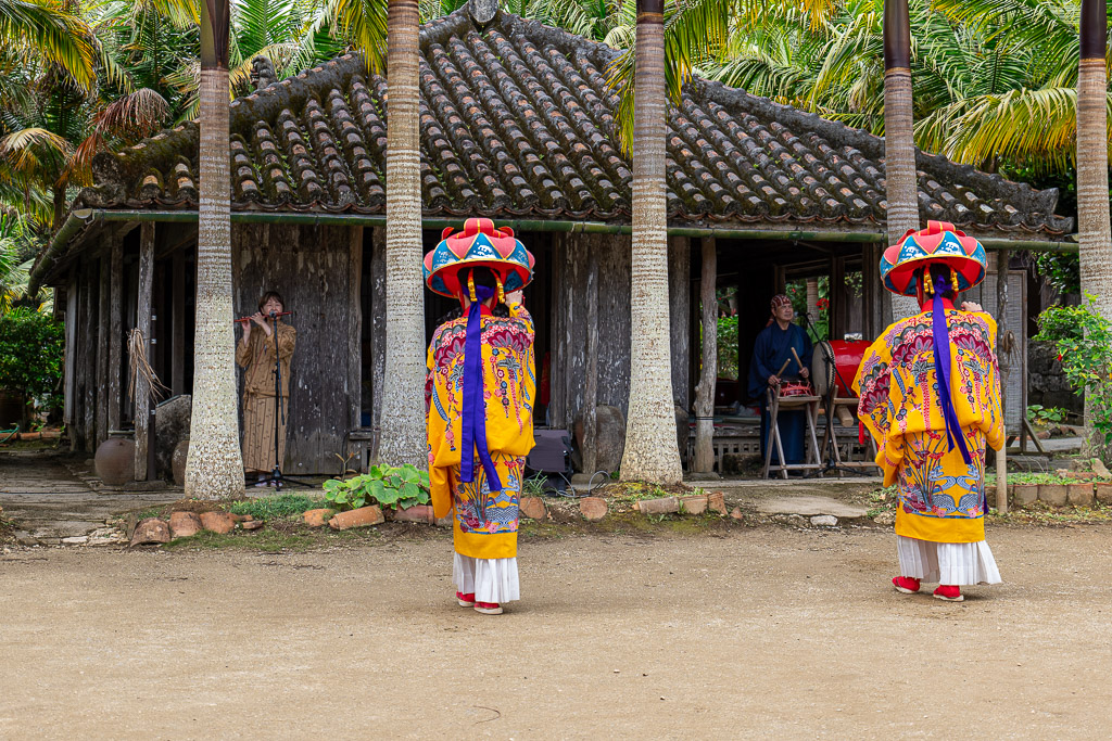Okinawan Hanagasa dance performance with two dancers in red-yellow flower hats and Bingata costumes in front of kominka house