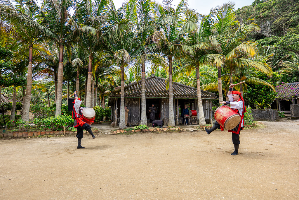 Eisa drum performance with two dancers swinging large drums and leaping in front of palm-tree-lined kominka houses