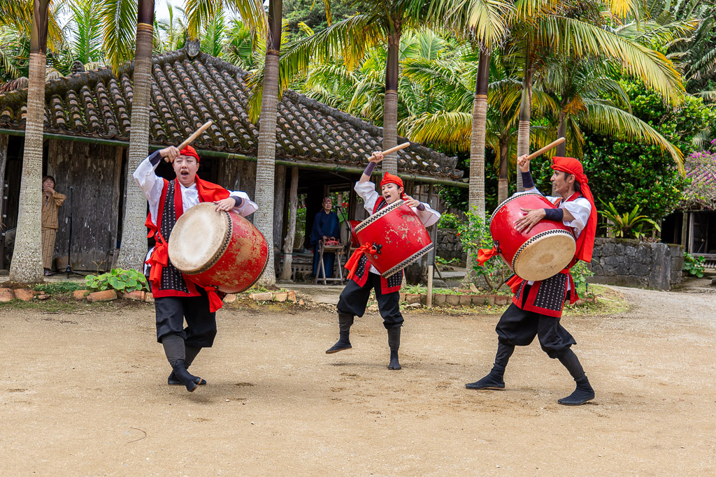 Eisa drum trio in red-and-black uniforms leaping under palm trees while striking large drums