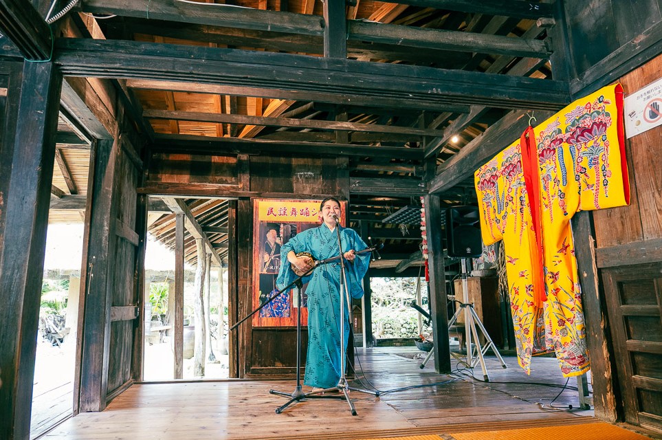 Sanshin workshop with instructor in Ryuso playing Sanshin in front of folk music dance posters inside kominka