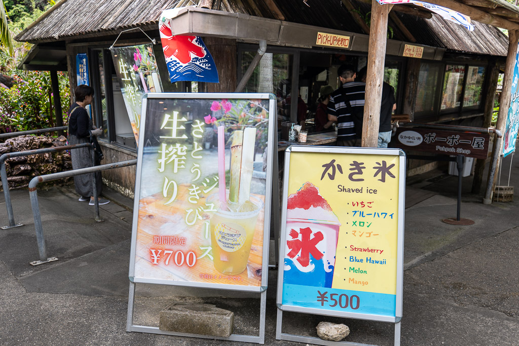 Pohpoh Shop exterior at Ryukyu Village with wooden signboard showing fresh sugarcane juice 700 yen and shaved ice 500 yen