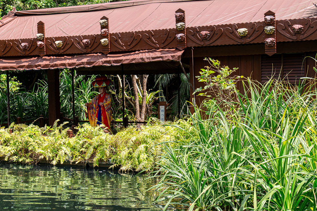 Lakeside Ryukyu-costume greeting stage at Bios no Oka — a staff member in traditional Ryukyu attire waves under a red-brown wooden canopy