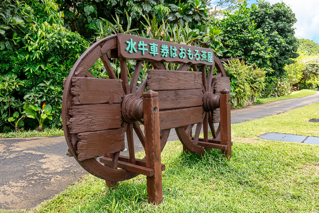 Water buffalo cart ticket signboard at Bios no Oka — a huge wooden cart wheel with a sign reading that tickets are sold at Omoro Chaya