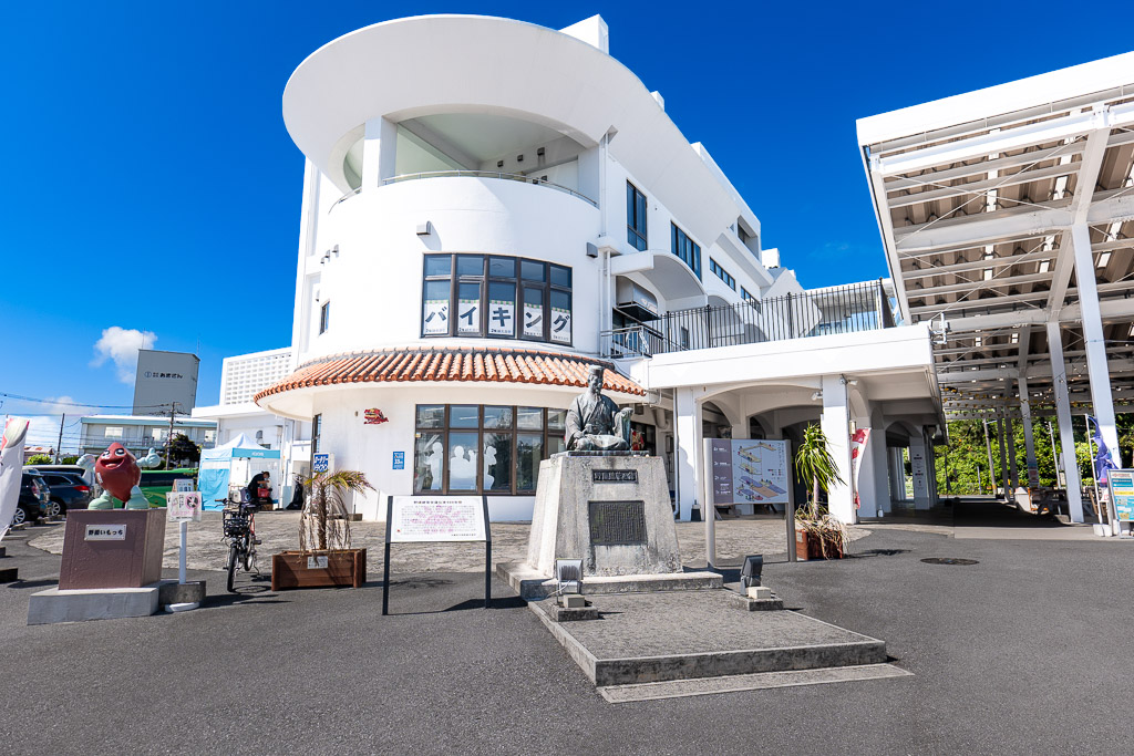 Round-fronted buffet building at Michi-no-Eki Kadena with the Noguni Sokan bronze statue and a sweet-potato mascot