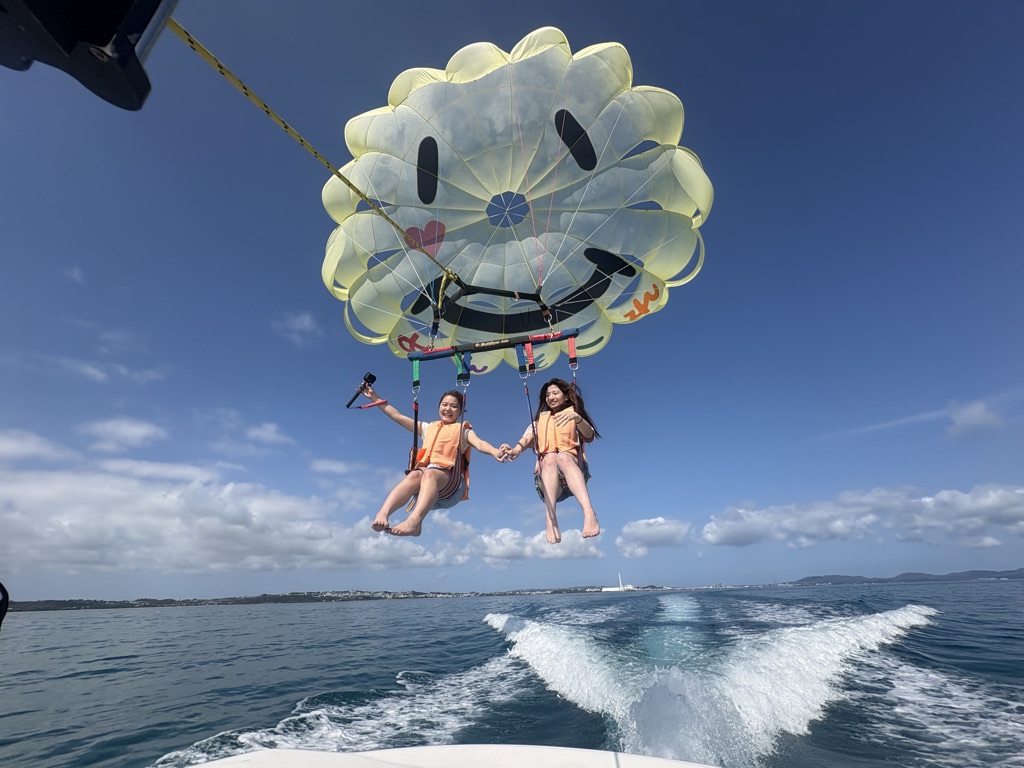 Two parasailers holding hands high above the ocean under a yellow smiley parachute in Okinawa