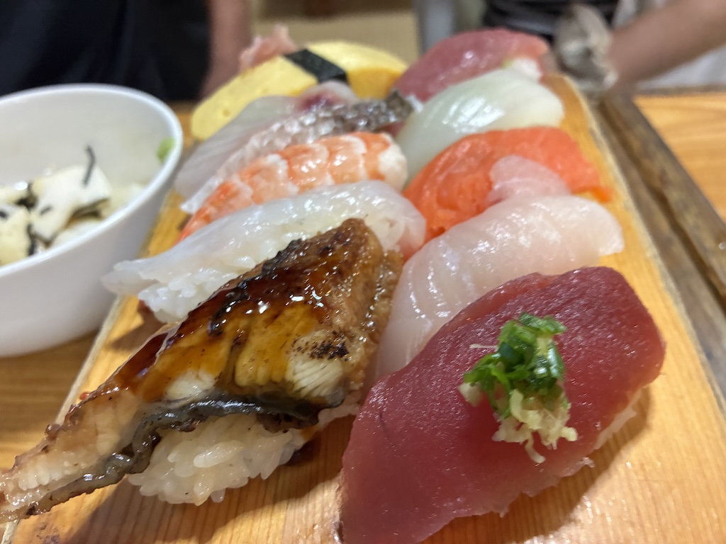 An overflowing wooden board of nigiri sushi — eel, tuna, shrimp, salmon, and tamago — at a traditional Japanese sushi counter
