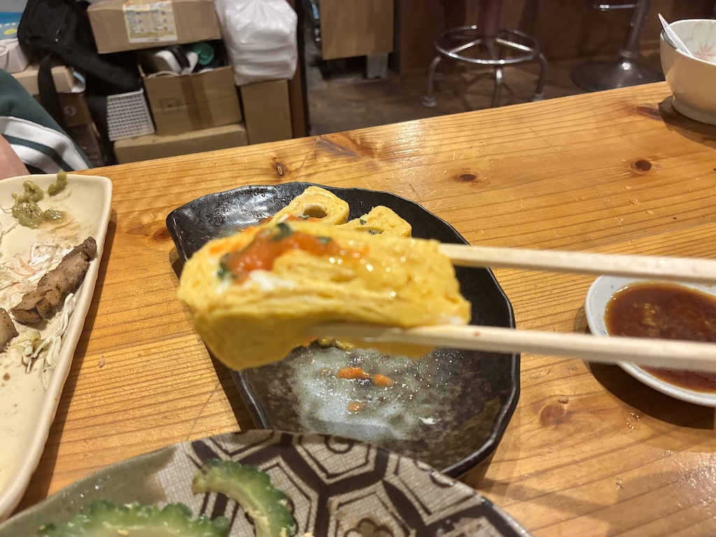 A rolled Japanese tamago omelette filled with red salted plum being lifted with chopsticks at an izakaya counter