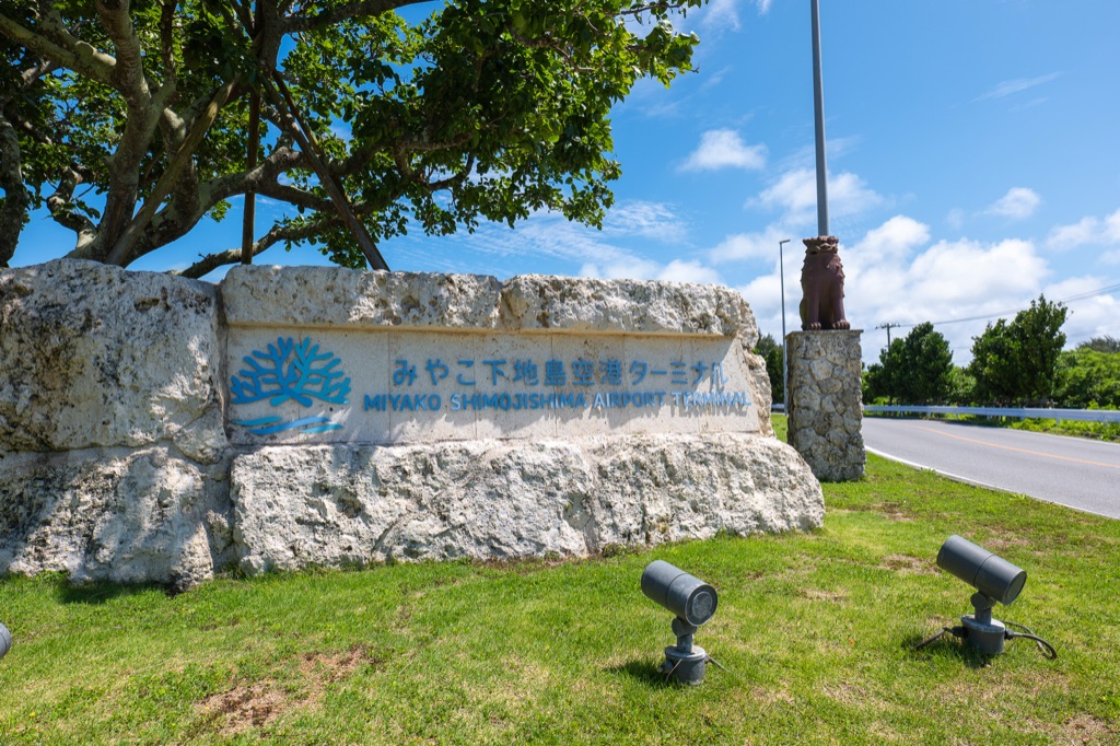 Welcome stone sign at Miyako Shimojishima Airport Terminal in Ryukyu limestone with sky and palms