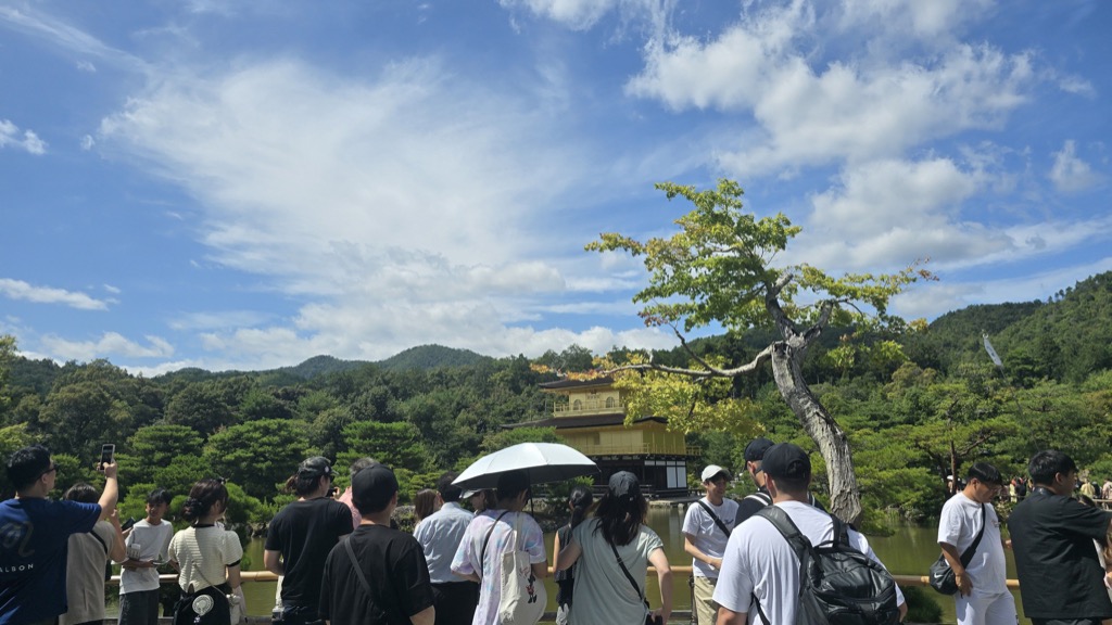 Visitors gathered at the front viewpoint of Kinkaku-ji Golden Pavilion with distant temple and lush summer greenery Kyoto