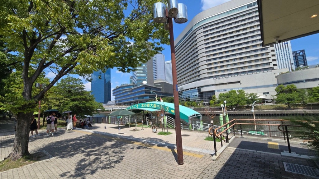 Pedestrians carrying sun parasols in the deep tree shade of a Nakanoshima riverside walkway under harsh early-September sun